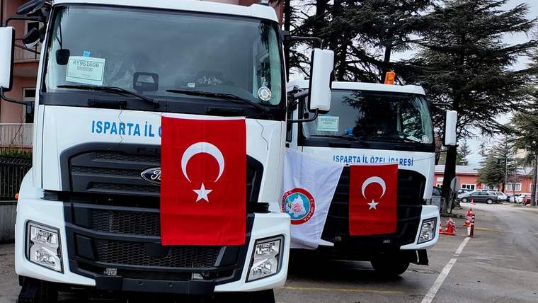 Türk bayrakları asılı, üzerinde Isparta İl Özel İdaresi yazan iki kamyon park alanında yan yana duruyor.