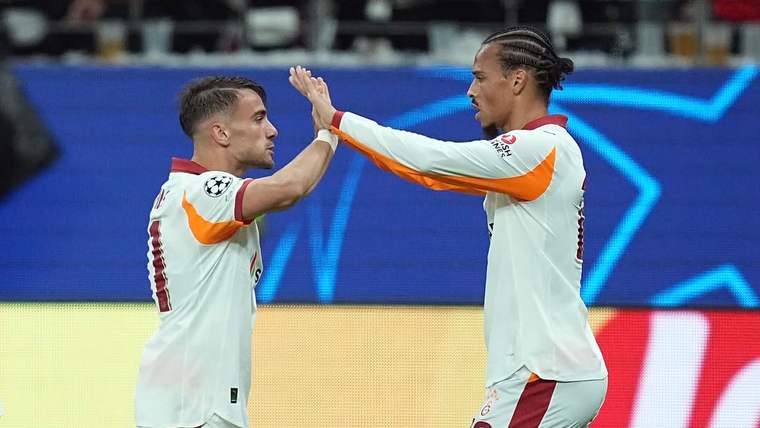 Saha kenarında beyaz formalı iki futbolcu, ellerini kaldırarak birbirine beşlik çakıyor.