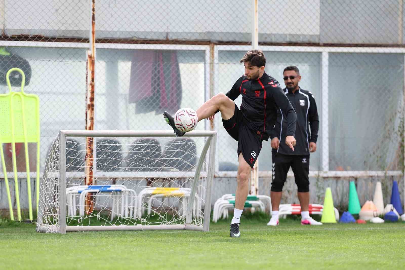 Siyah-kırmızı spor kıyafetli futbolcu, antrenman sahasında topa vururken arkasında bir kişi bekliyor.