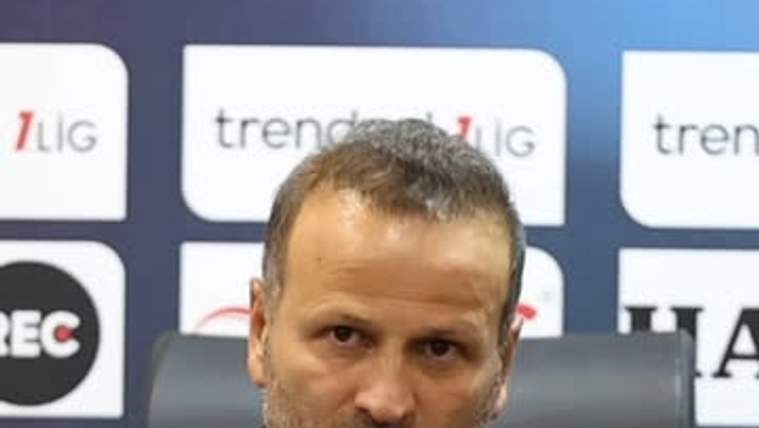 Mikrofonlar önünde oturan bir kişi, sponsor logolarının bulunduğu pano önünde basın açıklaması yapıyor.