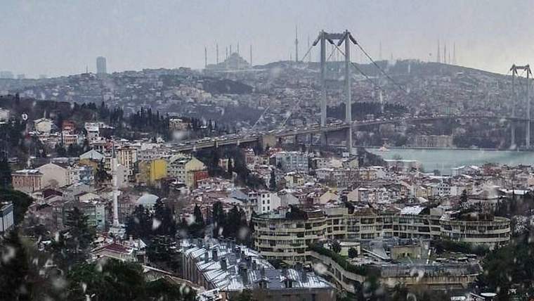 Kar yağışı altındaki yoğun yerleşim alanında, su üzerinde uzanan asma köprü ve çevredeki binalar görülüyor.