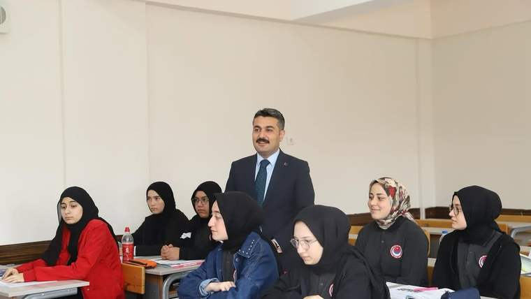 Sınıfta takım elbiseli bir erkek ve sıralarında oturan başörtülü öğrenciler görülüyor.