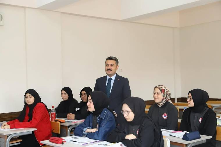 Sınıfta takım elbiseli bir erkek ve sıralarında oturan başörtülü öğrenciler görülüyor.