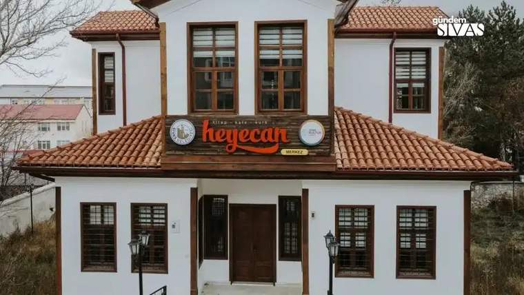Beyaz dış cepheli, ahşap detaylı bir bina, önünde "heyecan" yazılı tabela var.