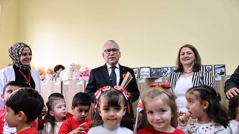 Bir odada çocuklar, üç yetişkin ve raflarda duran oyuncak bebekler birlikte görülüyor.