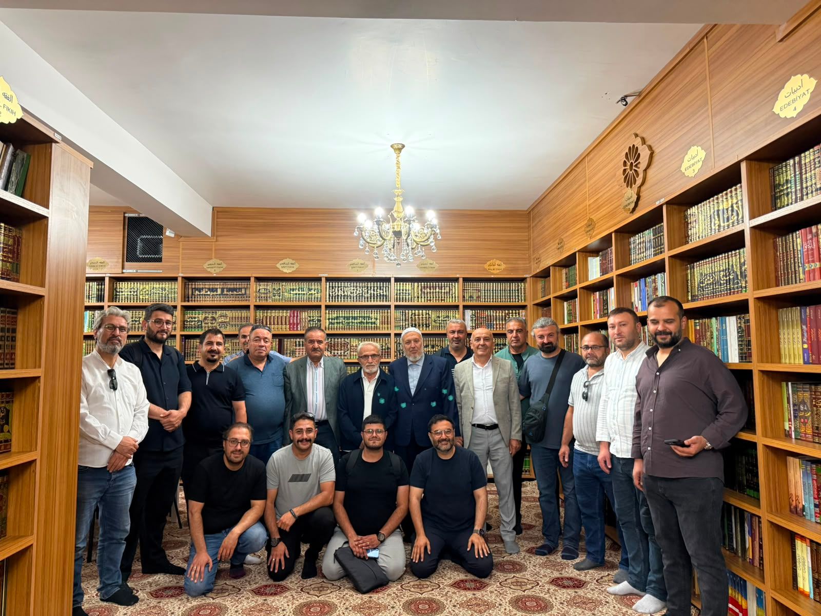 Kitaplıklarla çevrili süslü bir salonda, kalabalık bir erkek grubu ayakta ve önde oturarak poz veriyor.