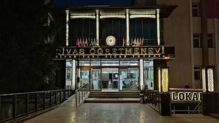 Gece aydınlatılan Sivas Öğretmenevi girişinde tabela, bayraklar ve rampalı yol görülüyor.