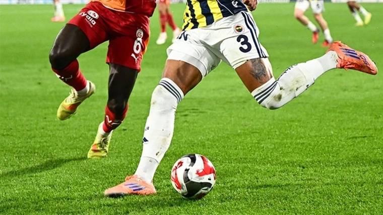 Futbol sahasında kırmızı ve sarı-lacivert formalı iki oyuncu topa doğru hamle yapıyor.