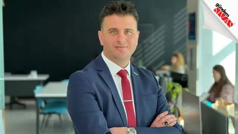 Takım elbiseli bir adam, kollarını bağlayarak modern görünümlü bir iç mekânda kameraya bakıyor.