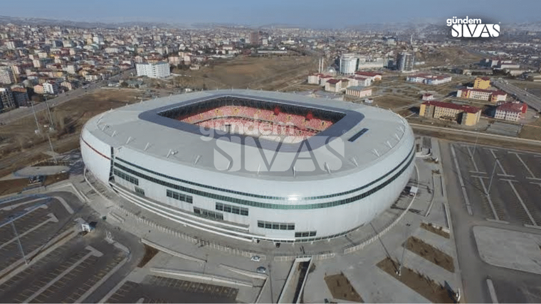 Modern bir stadyum, çevresinde park alanlarıyla birlikte görünmekte.