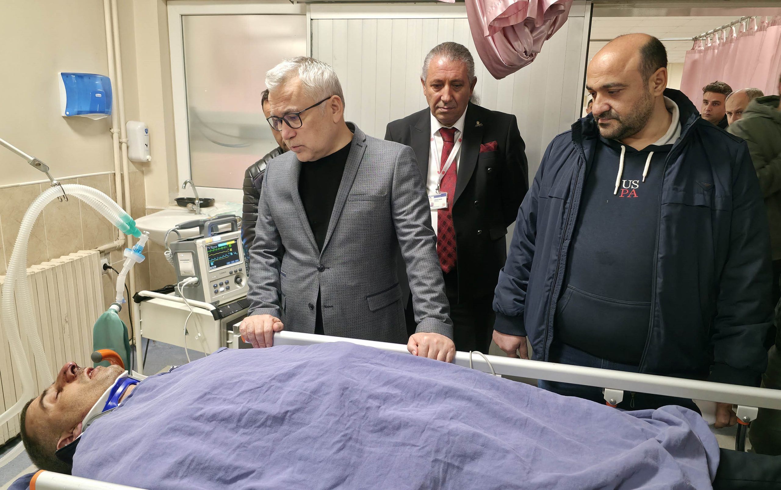 Hastanede yatan yaralı bir kişi, yanında üç kişi duruyor ve durumu inceliyor.
