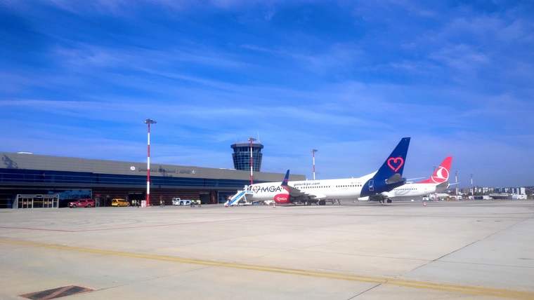 Havalimanı apronunda terminal binası ve kontrol kulesi önünde iki yolcu uçağı görülüyor.