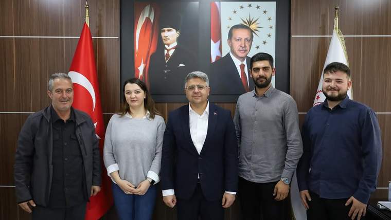 Türk bayrakları ve iki portre önünde, beş kişi bir makam odasında yan yana duruyor.
