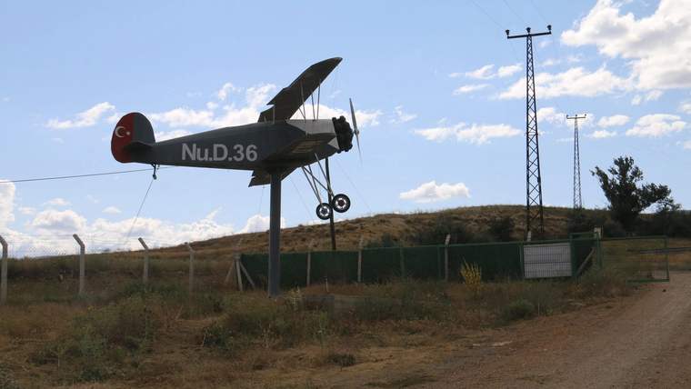 Açık havada, üzerinde “Nu.D.36” yazan pervaneli uçak maketi, çitlerle çevrili alanda sergileniyor.