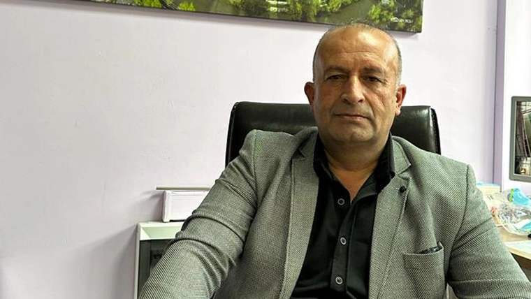 Koltukta oturan bir adam, gri ceket giymekte ve ciddi bir ifadeye sahip.