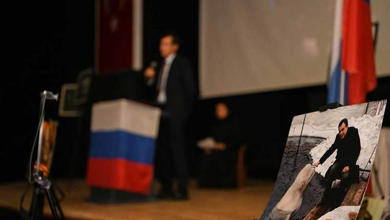 Bir sahnede, arka planda Türk ve Rus bayraklarıyla bir kişi konuşuyor, ön planda Andrey Karlov'un fotoğrafı yer alıyor.