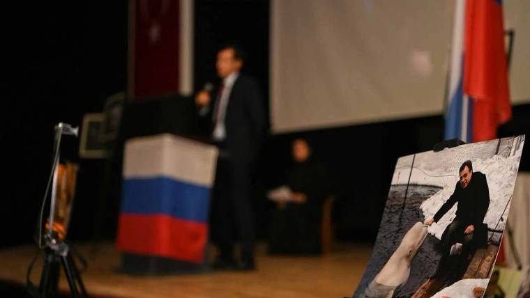Bir sahnede, arka planda Türk ve Rus bayraklarıyla bir kişi konuşuyor, ön planda Andrey Karlov'un fotoğrafı yer alıyor.