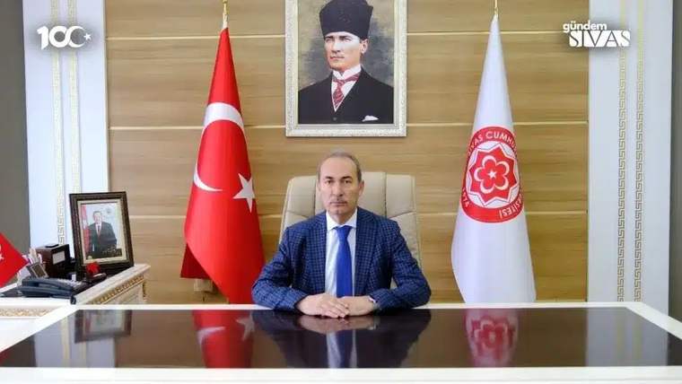 Bir masa başında oturan bir kişi, arkasında Türk bayrağı ve Sivas Cumhuriyet Üniversitesi bayrağı var.