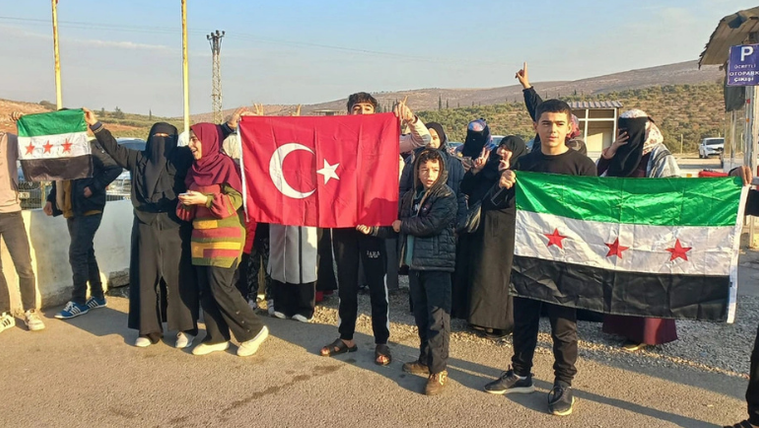 Yol kenarında toplanan bir grup, Türk bayrağı ile üzerinde yıldızlar bulunan Suriye bayraklarını taşıyor.