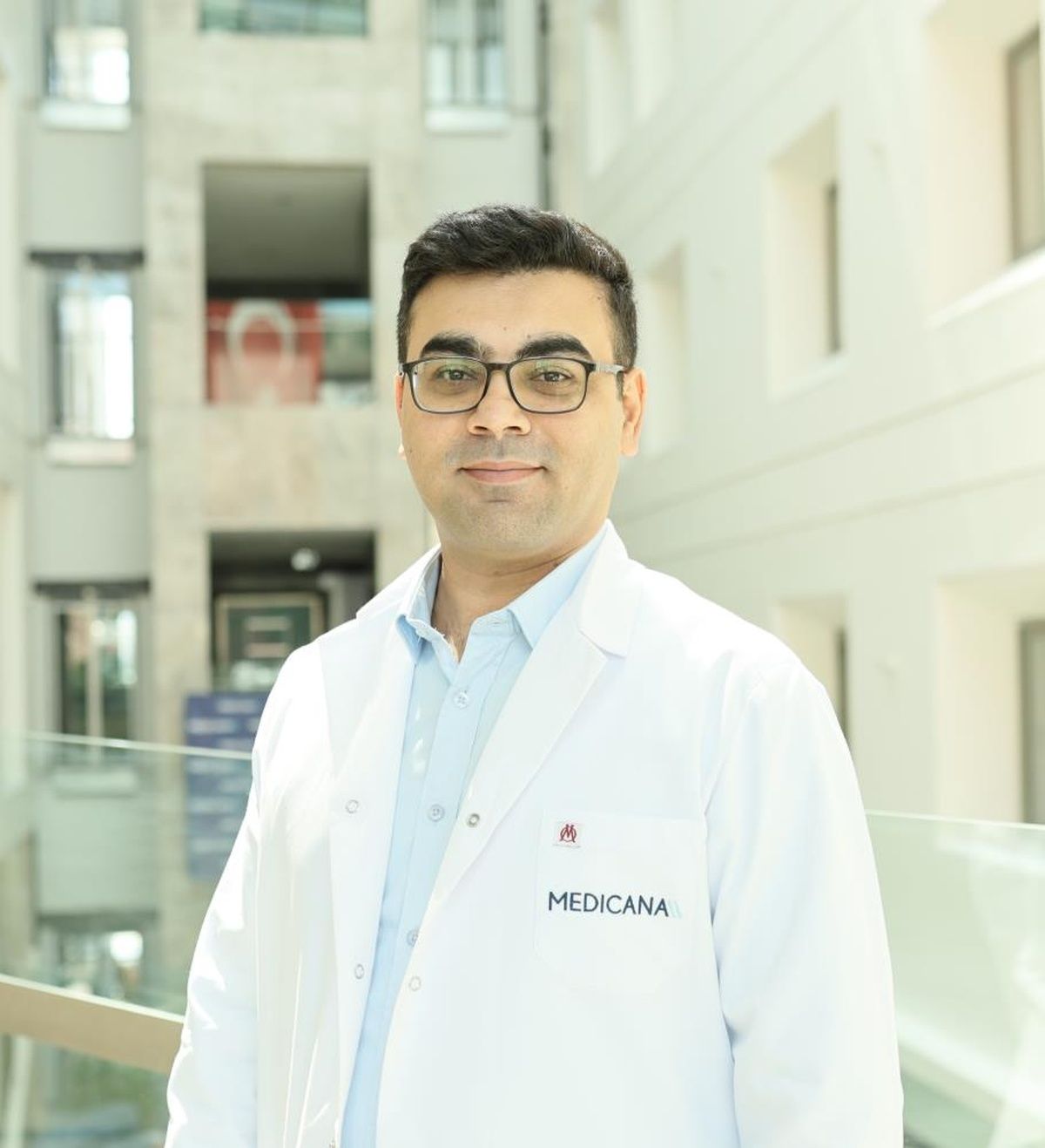 Gözlüklü bir kişi, üzerinde “MEDICANA” yazan beyaz önlükle aydınlık bir iç mekânda duruyor.