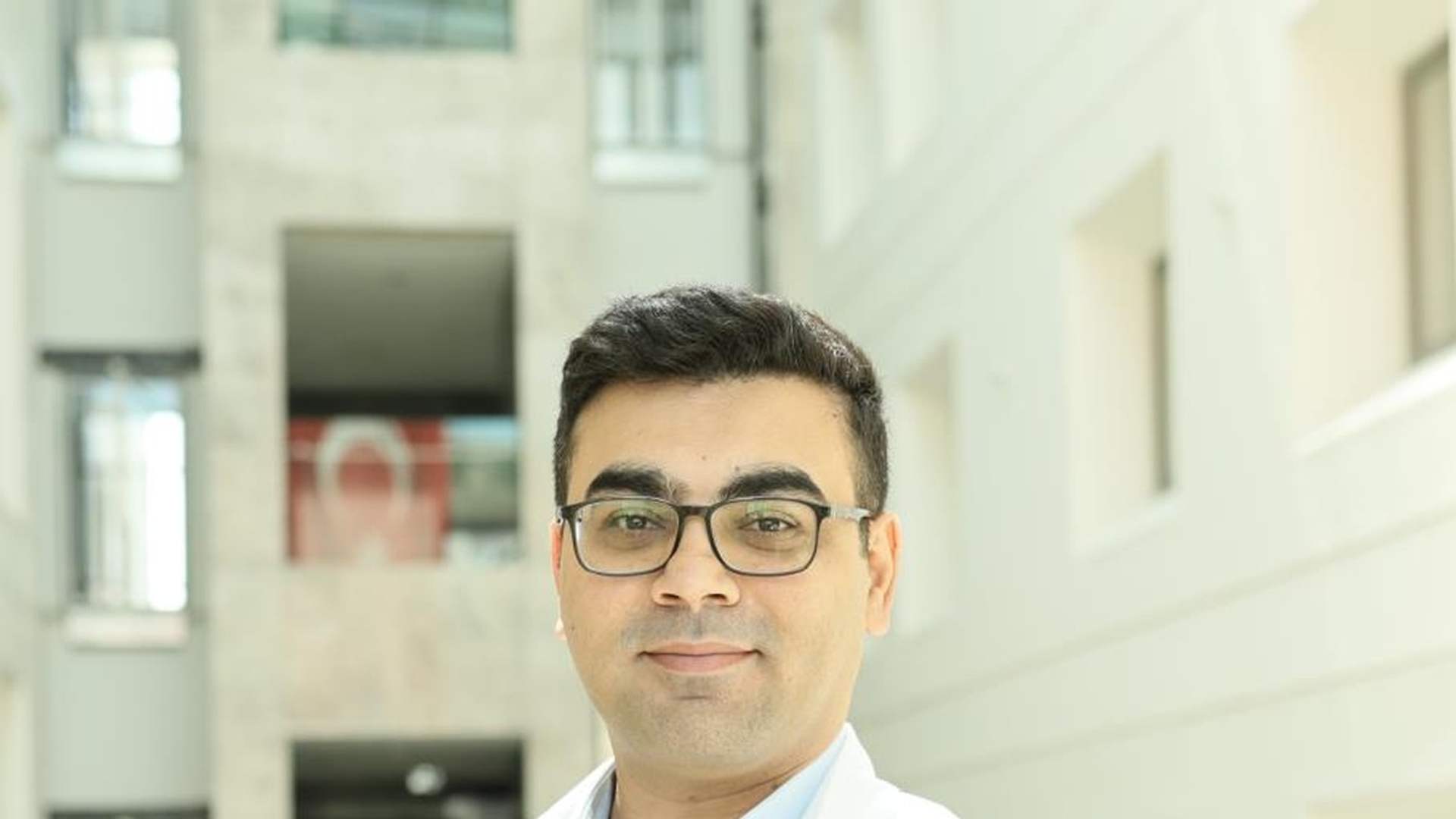 Gözlüklü bir kişi, üzerinde “MEDICANA” yazan beyaz önlükle aydınlık bir iç mekânda duruyor.