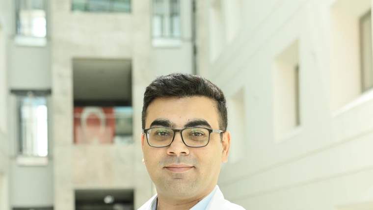 Gözlüklü bir kişi, üzerinde “MEDICANA” yazan beyaz önlükle aydınlık bir iç mekânda duruyor.