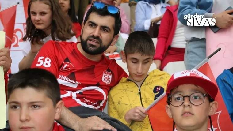 Kırmızı formalı bir adam, tribünde yanındaki sarı tişörtlü çocuğa doğru eğilirken çocuklar çevrelerinde oturuyor.