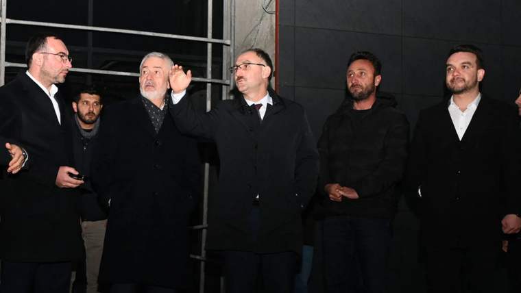 Bir grup erkek, bir inşaat alanında bir araya gelmiş, bir kişi elini kaldırarak konuşuyor.