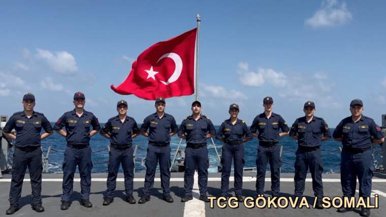 Denizci kıyafetleri giymiş on bir asker, Türk bayrağının önünde sıralanmış.