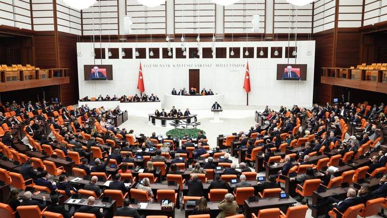 TBMM Genel Kurulu'nda milletvekilleri, oturumda yer alıyor; arka planda ekranlarda konuşan bir kişi görünüyor.