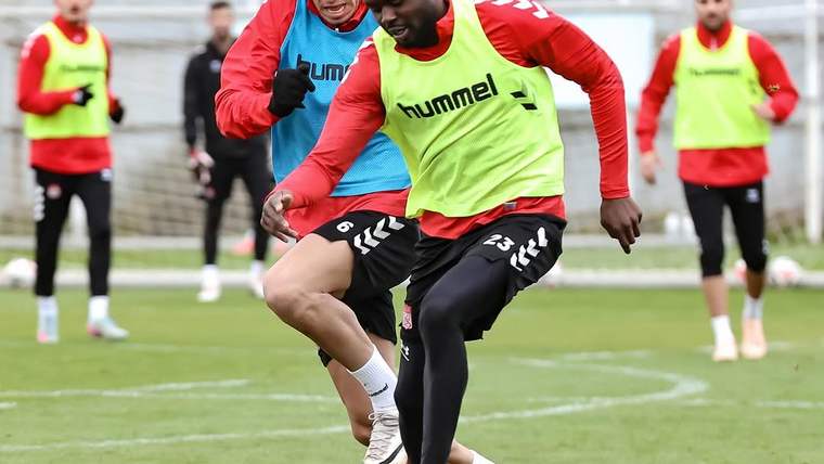 Yeşil sahada iki futbolcu top mücadelesi verirken, arkalarında antrenman yelekli oyuncular görülüyor.
