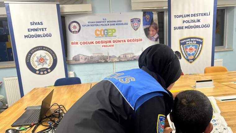 Mavi yelekli bir yetişkin, dizüstü bilgisayar ve kabloların bulunduğu masada bir çocuğun yanında duruyor.