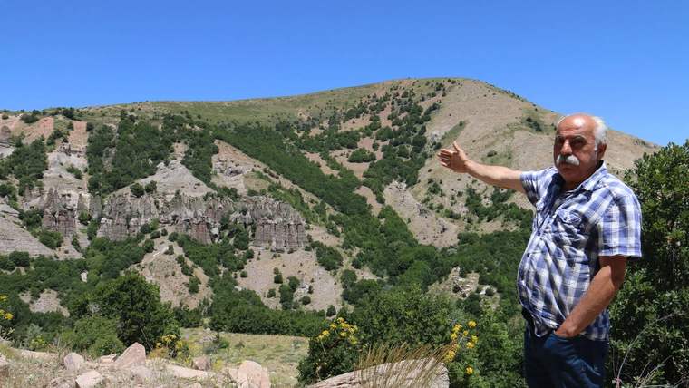 Bir kişi, arka planda dağlık bir alanı işaret ediyor. Yeşil ağaçlar ve açık mavi gökyüzü görünmekte.