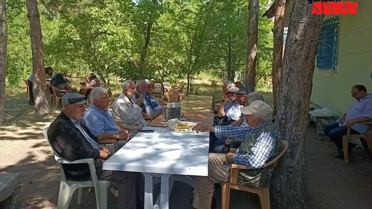 Ağaçların arasında oturan yaşlı erkekler, masada aşure ikramı yapılıyor.