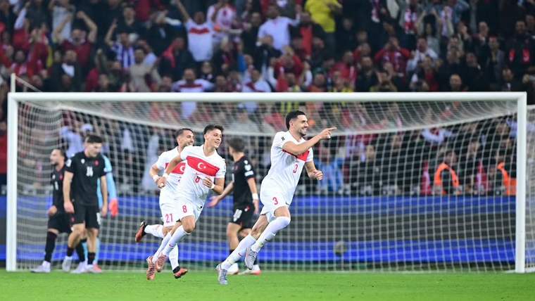 Beyaz formalı futbolcular, kalenin önünde sevinçle koşarken arkalarında tribünlerde taraftarlar görülüyor.
