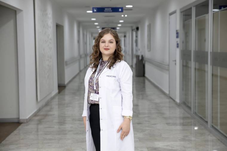 Hastane koridorunda beyaz önlüklü, boynunda kimlik kartı bulunan bir kadın ayakta duruyor.