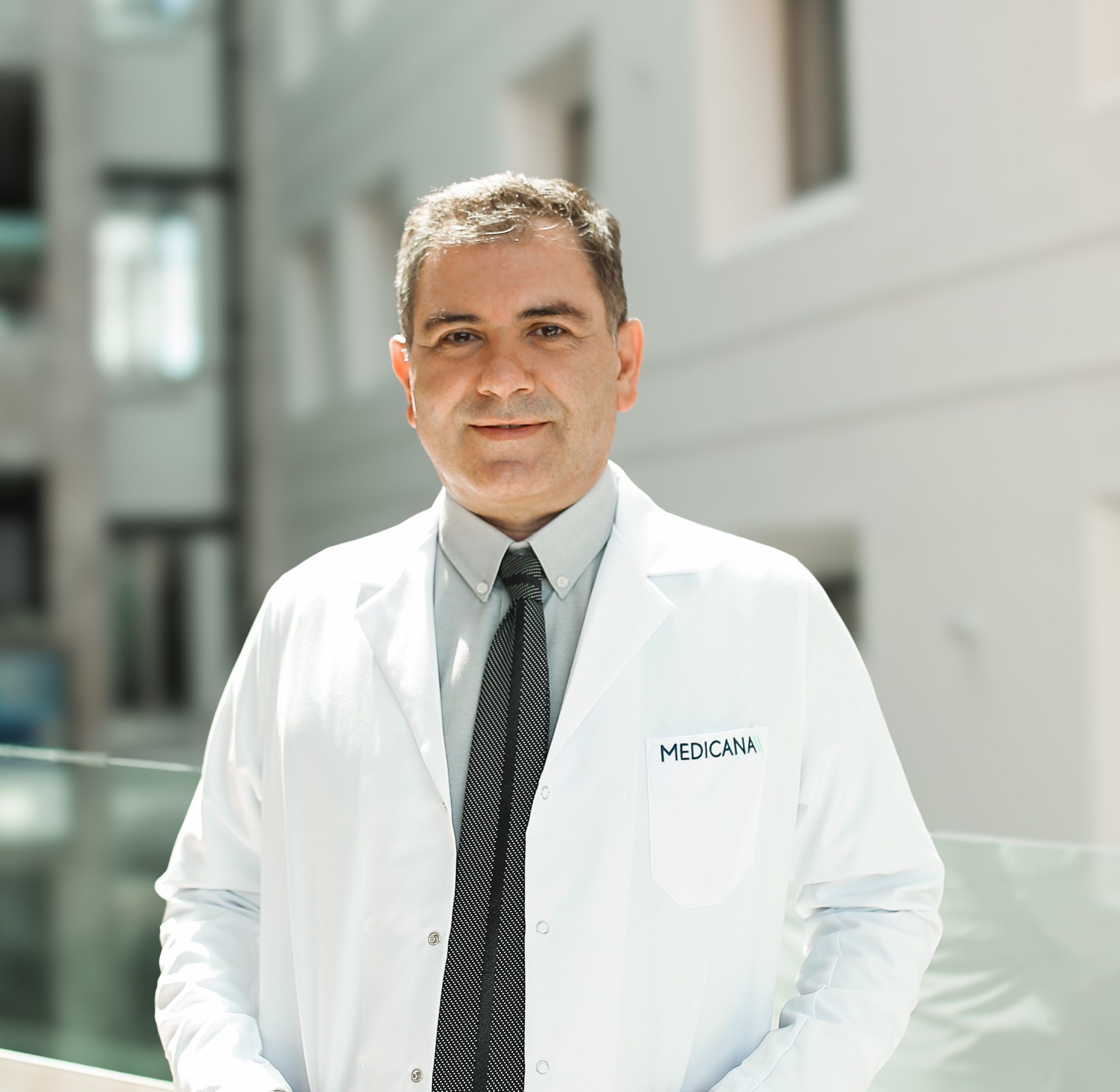 Beyaz lab coat giymiş bir doktor, arka planda modern bir bina ile gülümsüyor.