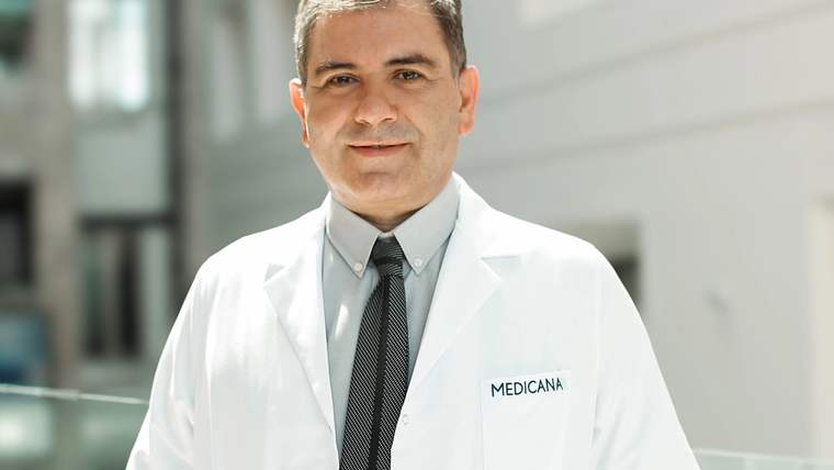 Beyaz lab coat giymiş bir doktor, arka planda modern bir bina ile gülümsüyor.