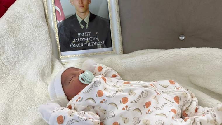 Beyaz battaniyeye sarılı emzikli bir bebek ve yanında çerçeve içinde üniformalı bir erkeğin portresi görülüyor.