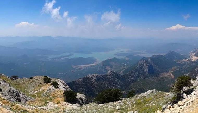 Kayalık dağ yamacından, uzakta göl ve dağlarla çevrili geniş bir manzara; mavi gökyüzünde bulutlar.