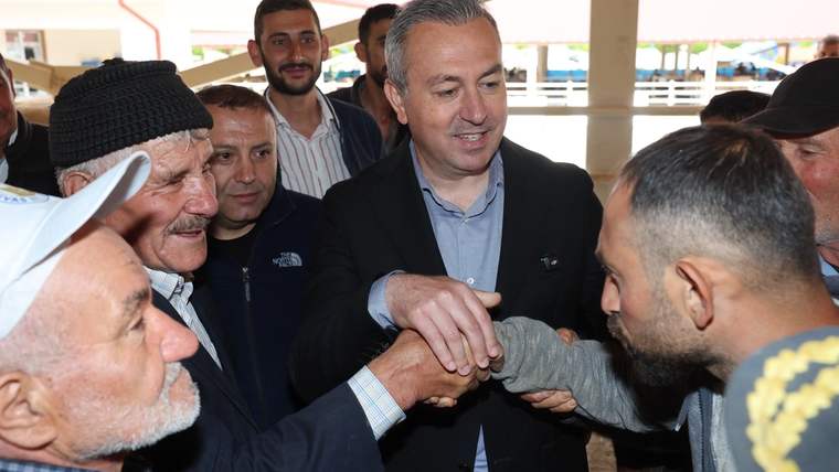 Bir grup insan, bir adamla el sıkışıyor; arka planda başka insanlar ve bir yapı görünmekte.