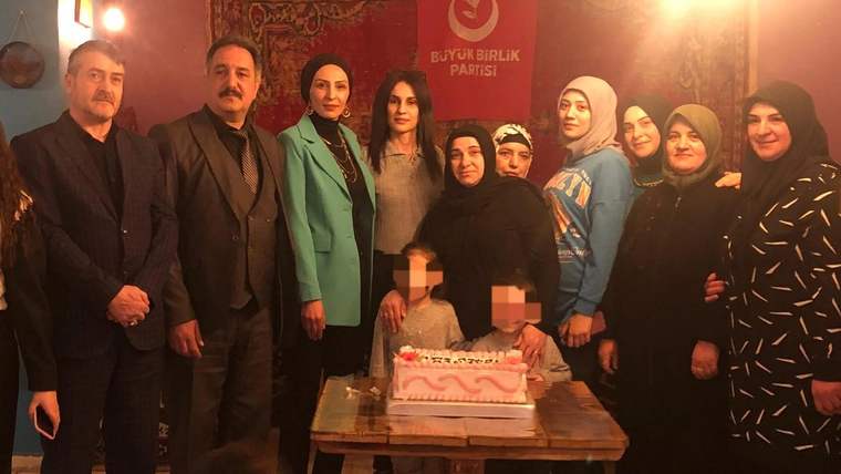 Bir odada, üzerinde Büyük Birlik Partisi yazılı pankart önünde duran yetişkinler ve iki çocuk, pastanın yanında poz veriyor.