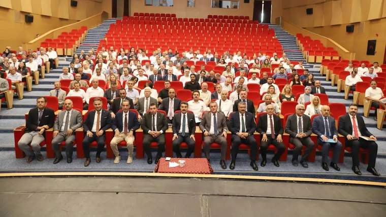 Ahşap panelli bir salonda, ön sırada oturan takım elbiseli kişiler ve arkalarında oturan kalabalık görülüyor.