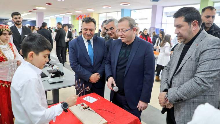 Bir grup insan, bir çocuğun yaptığı bir projeyi inceliyor; çocuk, masanın önünde duruyor.