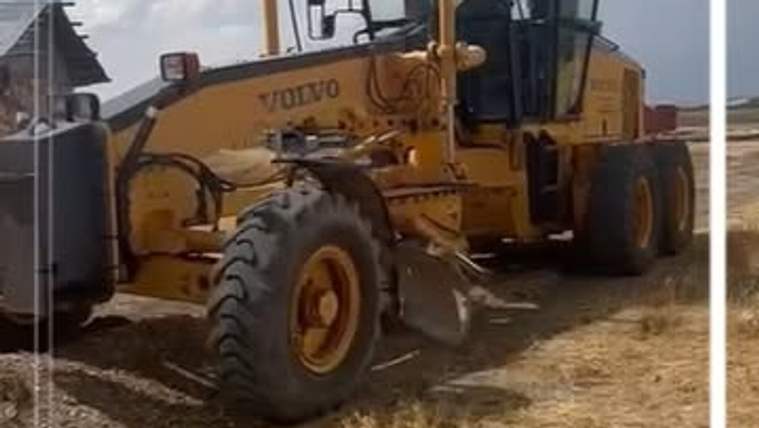 Sarı renkli VOLVO greyder, toprak zeminde bir evin yanında çalışırken görülüyor.