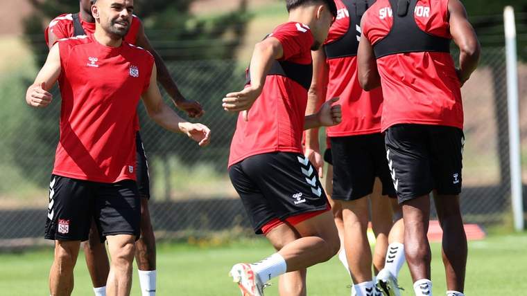 Kırmızı formalar ve siyah şortlar giyen futbolcular, çim sahada birlikte antrenman yapıyor.