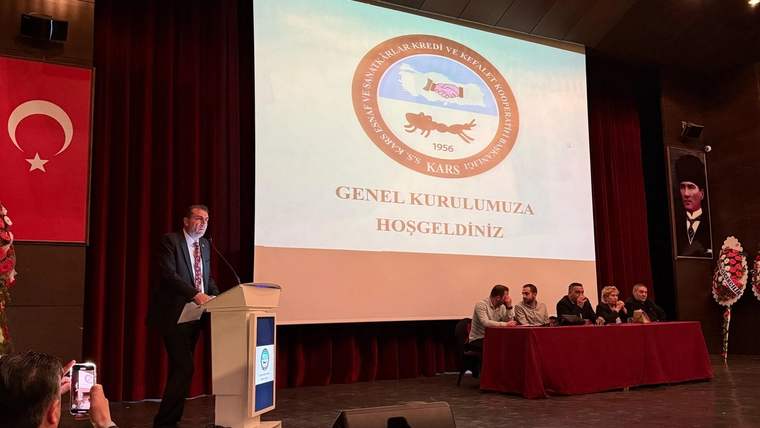 Sahne üzerinde bir kişi mikrofonla konuşuyor, arkasında genel kurul için hazırlanmış bir sunum var.