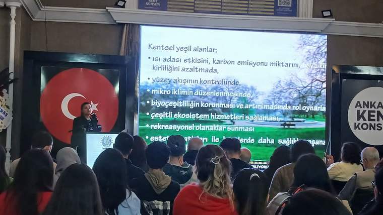 Bir salonda kürsüde konuşan kişi, çevresinde toplanan kalabalığa sunum yapıyor.