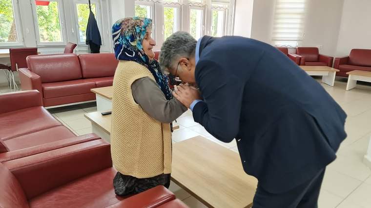 Bir kişi, huzurevinde oturan yaşlı bir kadının elini öpüyor, arka planda kırmızı koltuklar var.