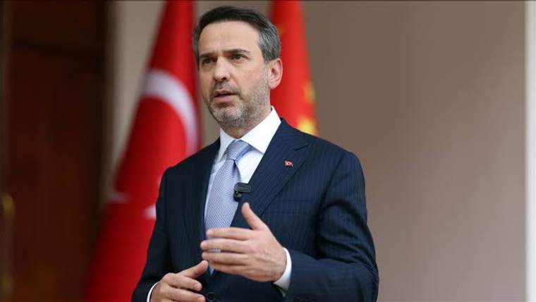 Takım elbiseli bir kişi konuşurken el hareketi yapıyor; arka planda Türk bayrağı görülüyor.